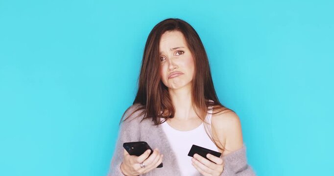 Sad young woman facing online payment problem with credit card and smartphone, looking disappointed and making sorry facial expression in cozy grey cardigan isolated on vibrant blue background