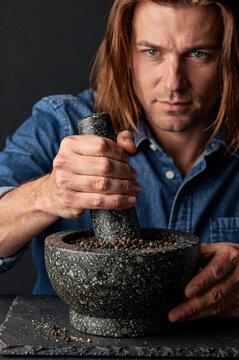 Man crushes black peppercorns inside a granite mortar using a heavy pestle. He wears a blue denim shirt against a dark studio setting with focused side lighting on his hands.