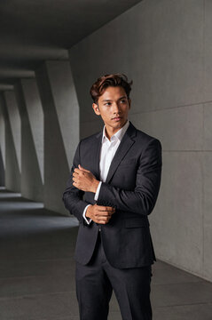 Man in dark charcoal suit adjusts his sleeve cuff while standing in a concrete corridor. Hard side lighting creates high contrast shadows against the minimalist gray pillars and floor.