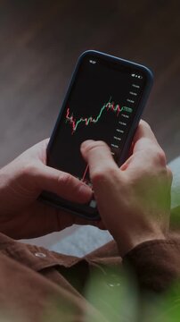Close-up of a trader hands holding a smartphone displaying a cryptocurrency candlestick chart. Investor tracks stock market or crypto data and exchange rates on a mobile trading app. Vertical video