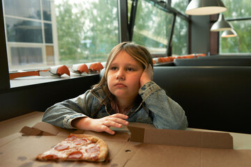 A primary school-aged girl sits at a table in a pizzeria. A remnant of pizza lies on the table in front of her. Paper masks of large-beaked birds sit on the windowsill.  © ROMAN DZIUBALO