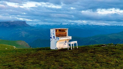 Dzhily Su, Russia - June 4, 2026: Old piano standing outdoors with mountains in the background, creating an unusual scene with a weathered instrument, highland landscape, green hills, and scenic Cauca © batkanov