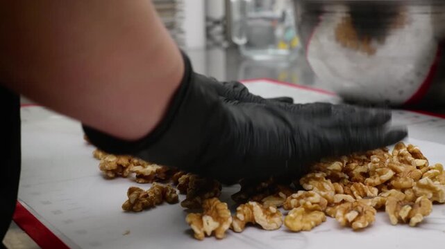 A professional chef or home baker wearing black protective gloves carefully arranges a heap of raw, peeled walnut kernels on a white silicone baking mat. This footage captures the essential steps of p