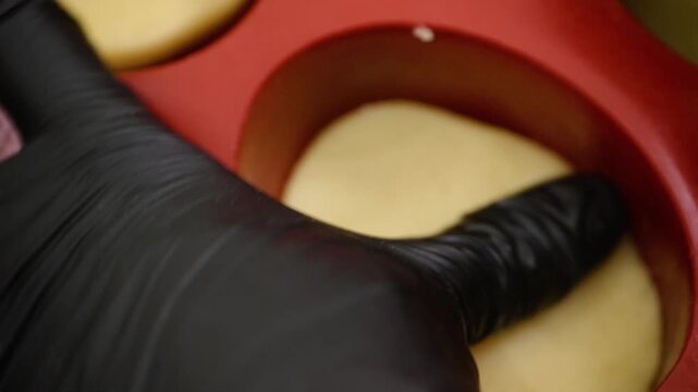 High-angle, close-up shot of a professional pastry chef wearing black nitrile gloves carefully shaping raw pastry dough into a round red silicone mold. This footage captures the meticulous process of 