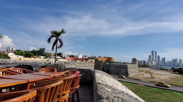 Beautiful view from the stone defensive walls showcasing empty restaurant tables a palm tree and the modern skyline of Cartagena Colombia
