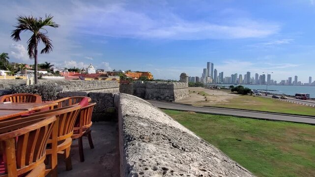 Beautiful view from the stone defensive walls showcasing empty restaurant tables a palm tree and the modern skyline of Cartagena Colombia