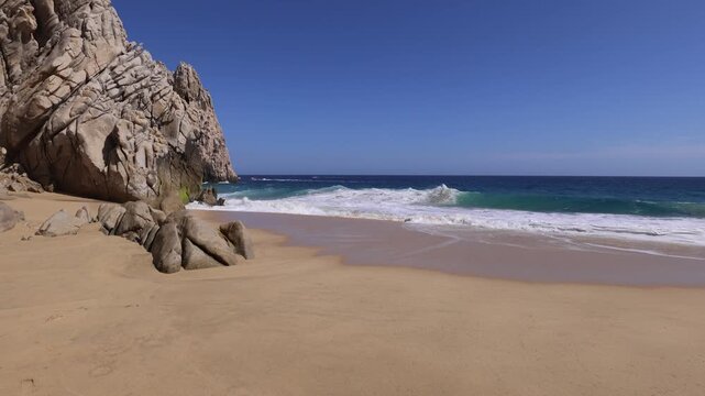 Scenic view of powerful ocean waves breaking on a golden sand beach next to dramatic rock cliffs in Cabo San Lucas Mexico at Los Arcos