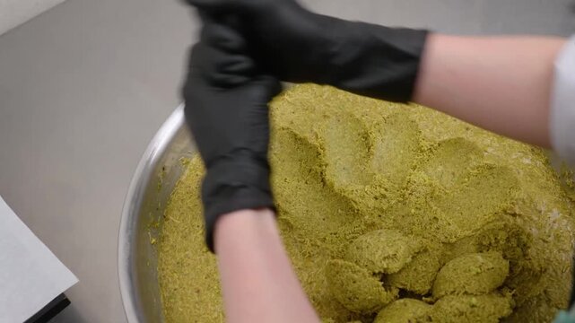 Close-up of a baker in black nitrile gloves portioning a textured green pistachio and kataifi mixture with a metal scoop in a professional stainless steel kitchen. Ideal for culinary, dessert trends, 
