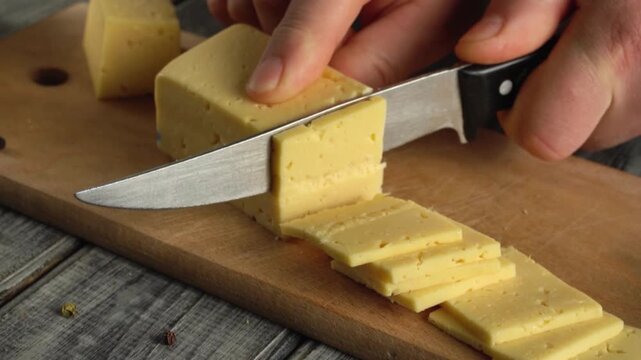 Hand slicing block of yellow cheese into even pieces on wooden cutting board with rustic background and kitchen elements visible