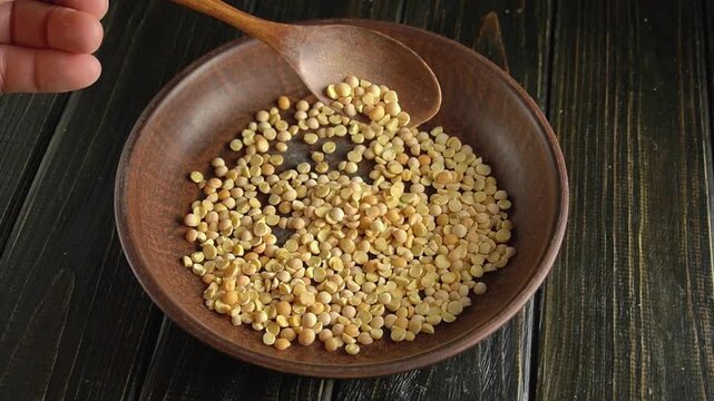 Hand with wooden spoon scooping yellow split peas from a brown bowl on a dark wooden surface, showcasing the gradual pouring action
