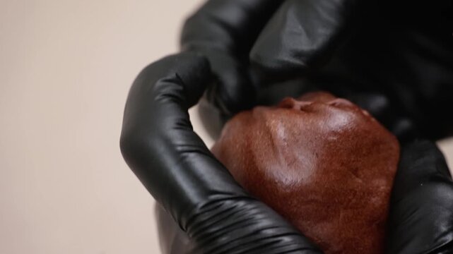 High-angle closeup of a professional pastry chef wearing black nitrile gloves carefully shaping or filled a round, golden-brown dessert pastry, possibly a viral Dubai chewy cookie or stuffed pie, on a