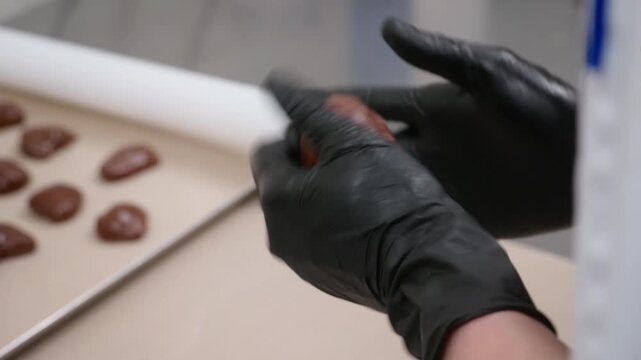 Professional pastry chef wearing black nitrile gloves manually rolling and shaping portions of chocolate dough into uniform balls on a parchment-lined baking sheet. This high-quality footage showcases