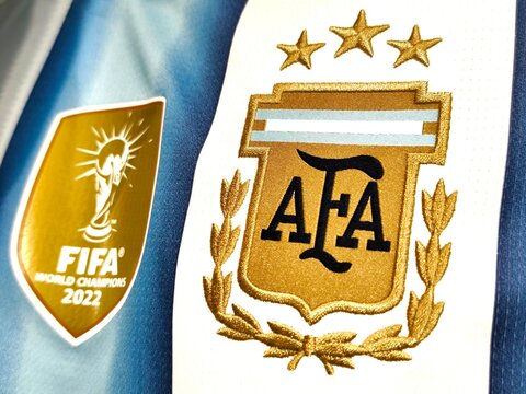 Closeup view of the official Argentina home jersey, showcasing the golden three-star AFA crest and the FIFA World Cup champions badge against the iconic blue and white stripes.