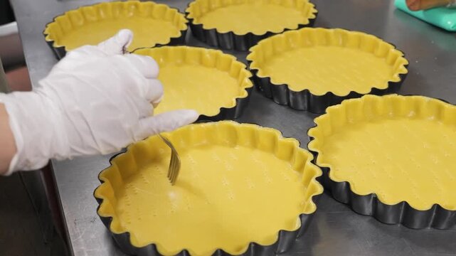 A close-up shot of a baker wearing white gloves using a fork to "dock" or prick holes in raw shortcrust pastry dough inside fluted tart pans. This professional technique allows steam to escape during 