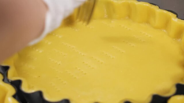 Professional baker using the tines of a metal fork to dock raw shortcrust pastry dough in a fluted tart pan. This essential baking technique, known as docking, prevents air bubbles from forming and en