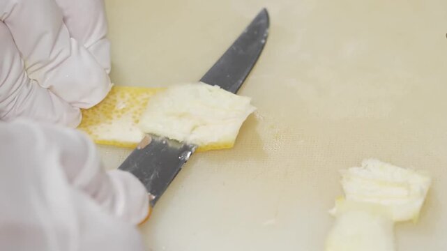 A close-up shot of a chef's hands in white gloves using a sharp knife to skillfully remove the yellow zest from a fresh lemon on a clean cutting board. This high-quality footage captures the detailed 