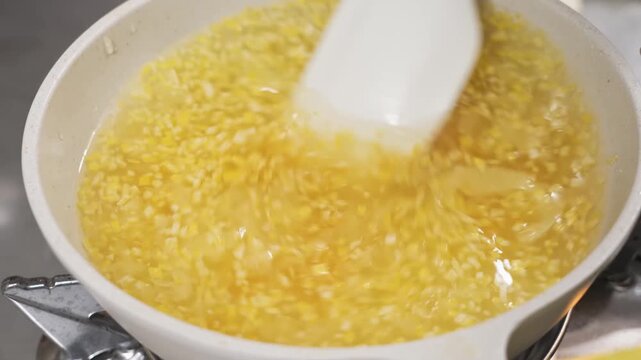 close-up shot of a thick, yellow sweet dessert pudding or pie filling being stirred with a white spatula in a white ceramic pan. The mixture features a bright citrus hue with visible small granules, c