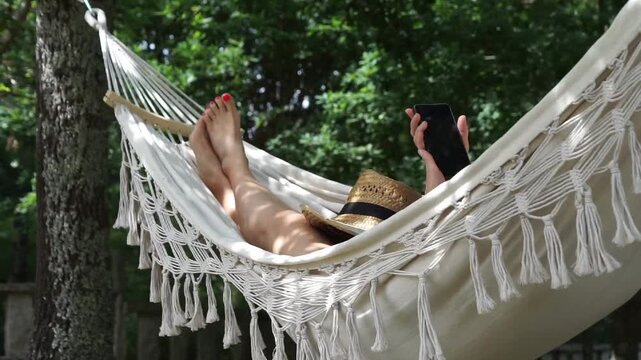 Low angle shot of a woman's legs resting in a white fringe hammock outdoors, as she raises her hand to check her mobile phone on a sunny day.