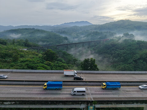 Aerial view of Jembatan Kereta Api Cikubang railway bridge spanning a lush valley with morning mist, near a highway with traffic in Kecamatan Cipatat, Jawa Barat, Indonesia.