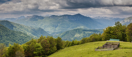 Obraz premium Green hills and dense forests extending to distant mountains under a cloudy sky, featuring an old wooden fence structure