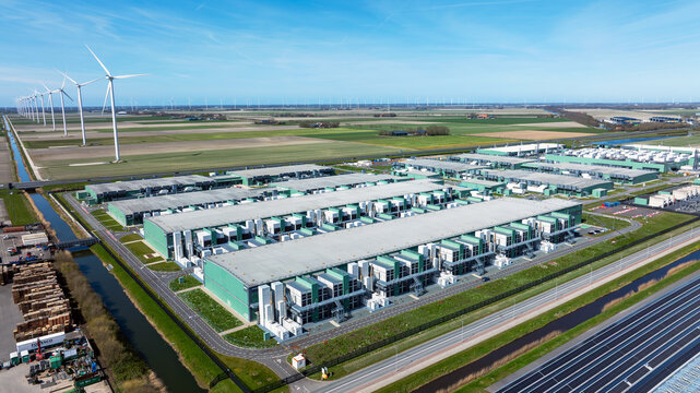 Aerial view of a large data center facility with industrial buildings surrounded by green fields and wind turbines in Middenmeer, Noord Holland, Netherlands.