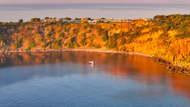 Aerial view of the rugged coastline of Pollina, featuring golden cliffs at sunset with a small boat floating in the calm turquoise sea below, Pollina, Sicilia, Italy.