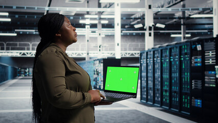 Fototapeta premium Data center administrator uses green screen laptop in a data center to inspect hardware and maintain system stability.