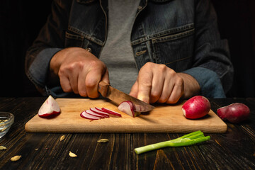 A cook slices red radishes and green onions on a wooden cutting board. The setting is a kitchen with dim lighting. The focus is on the cutting process with vegetables nearby © Віталій Б.
