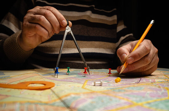 A person holds a compass with one hand and a pencil with the other while planning a route on a map. Push pins are placed at different points on the map