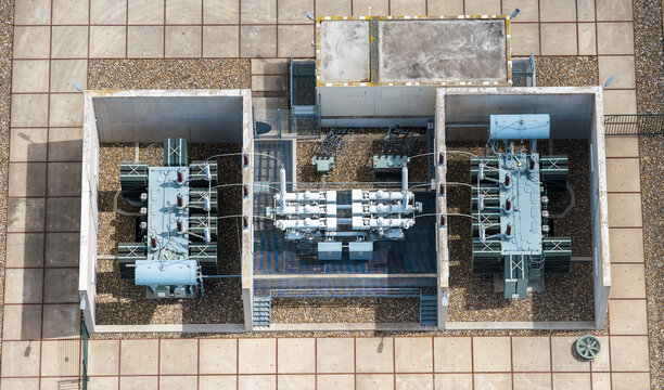 Aerial view of an electrical substation with industrial transformers and switchgear equipment housed within concrete enclosures on a paved site in Espel, Flevoland, Netherlands.