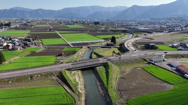 Green fields stretch under clear blue sky. River winds through farmland, crossing a road. Mountains frame the distant horizon. Village rooftops dot the landscape below