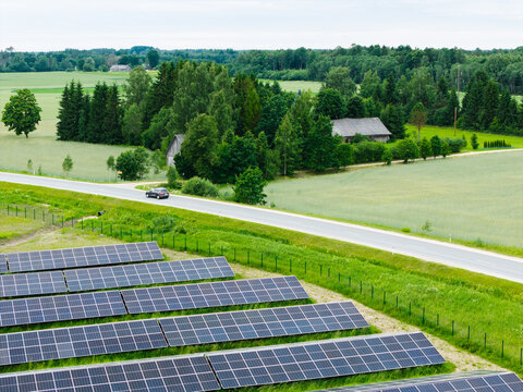 Aerial view of a solar farm situated in a rural landscape with green fields, dense forest, and a road with a car passing by in Suntazi, Ogre Municipality, Latvia.
