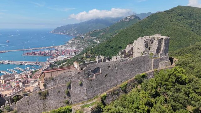 Aerial view of Castello di Arechi medieval fortress on lush forested hill with Salerno port, marina, and Amalfi Coast stretching along the horizon, Italy