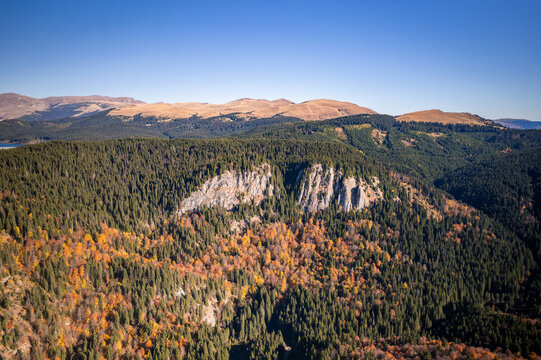 Aerial view of Lacul Scropoasa surrounded by dense autumn forests with vibrant foliage and rocky cliffs under a clear blue sky in Scropoasa, Judetul Dambovita, Romania.