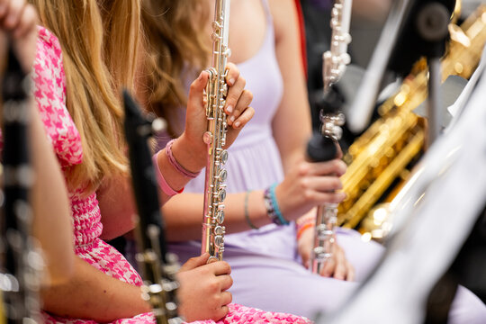 Young flute players hold their silver instruments upright during a school concert band performance, with the gold curve of a saxophone section visible just behind them. 