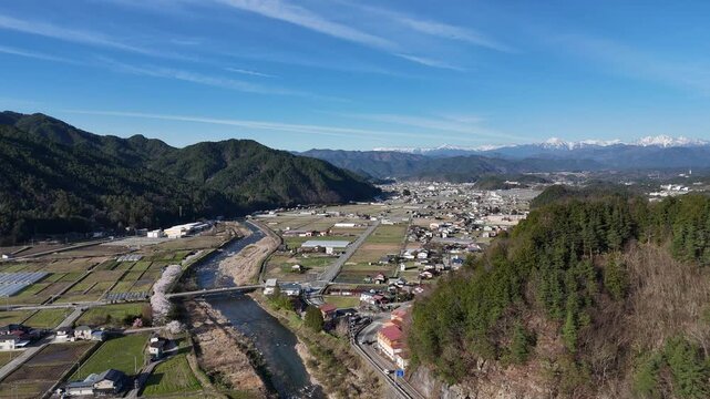Blue sky above winding river through green hills. Village nestled between forest and farmland below. Snow-capped mountains frame distant horizon. Serene landscape with no people visible