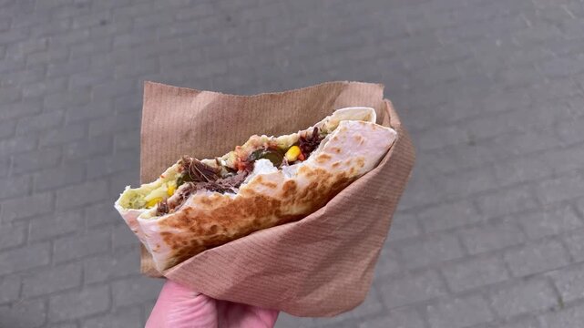 Grilled tortilla roll with meat filling in a paper bag, street food against a city sidewalk background, Mexican dish, street food