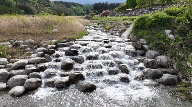 Clear water flows over smooth stones. Rustic stream rushes through mossy banks. Lush green hills frame the scene. Nature's quiet energy is evident. Ideal for serene landscapes or rural settings