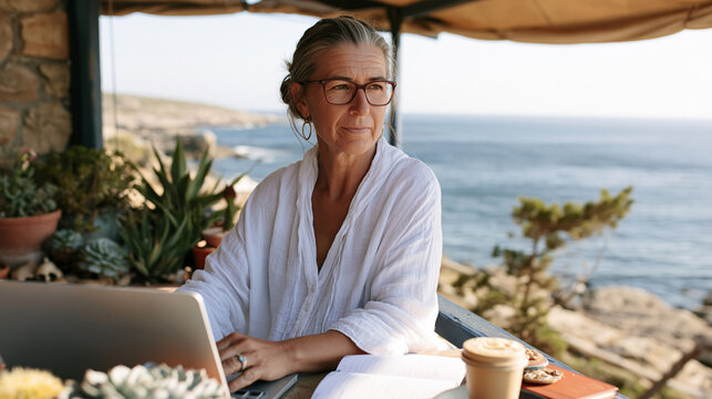 Mature woman working remotely on laptop at coastal retreat, enjoying tranquil sea view from balcony