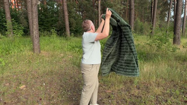 A mature woman shakes out a dirty picnic blanket in a summer forest, outdoor recreation and a picnic in nature
