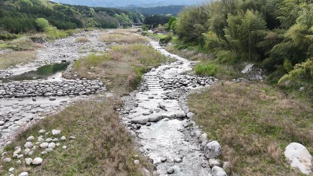 Riverside stream flows gently through stone bed. Grassy banks stretch wide, lined with green trees. Sunlight filters through foliage, casting soft shadows. No people, no animals