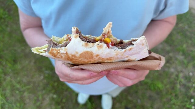 A woman holds a half-eaten grilled meat wrap outdoors, a close-up of street-food shawarma in a park
