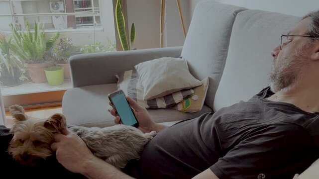 Man with glasses chatting on his smartphone and petting his dog.Pet on its owner's lap receiving affection .Horizontal 4k footage. Relaxing moments. Pet lovers. Technology at home