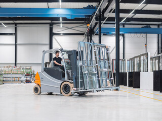 Fototapeta premium Forklift driver moves an A-frame rack of insulated glass through a glazing warehouse, the cab seen in near profile.