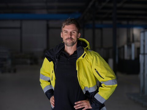 Worker stands with hands on hips in a hi-vis jacket, a slight smile and assertive stance in a dim warehouse.
