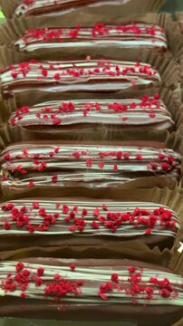 Close-up glossy chocolate eclairs lined in paper cups topped with white drizzle and red crumble, rows of decadent pastries