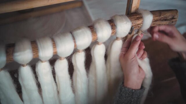 Close-up of artisan woman's hands tying knots with cord on a macrame tapestry made with wool roving, showcasing handmade textile art and creative craft process.
