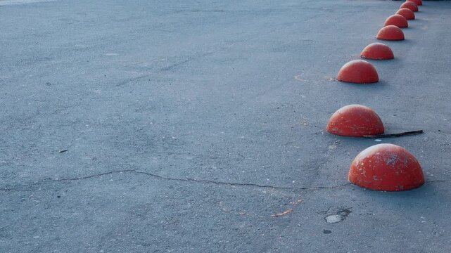 Red hemispheres on asphalt pedestrian path, row of weathered orange bollards creating rhythmic barrier in empty urban pedestrian