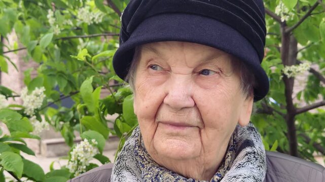 Elderly woman in park close-up lilac, contemplative gaze away from camera, black hat and patterned scarf, spring foliage and lilac