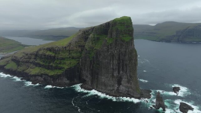 Aerial drone shot panning left, revealing the majestic Ei&eth;iskollur mountain and its dramatic sea cliffs on the Faroe Islands. Overlooking the village of Ei&eth;i and the rugged coast with crashing waves.